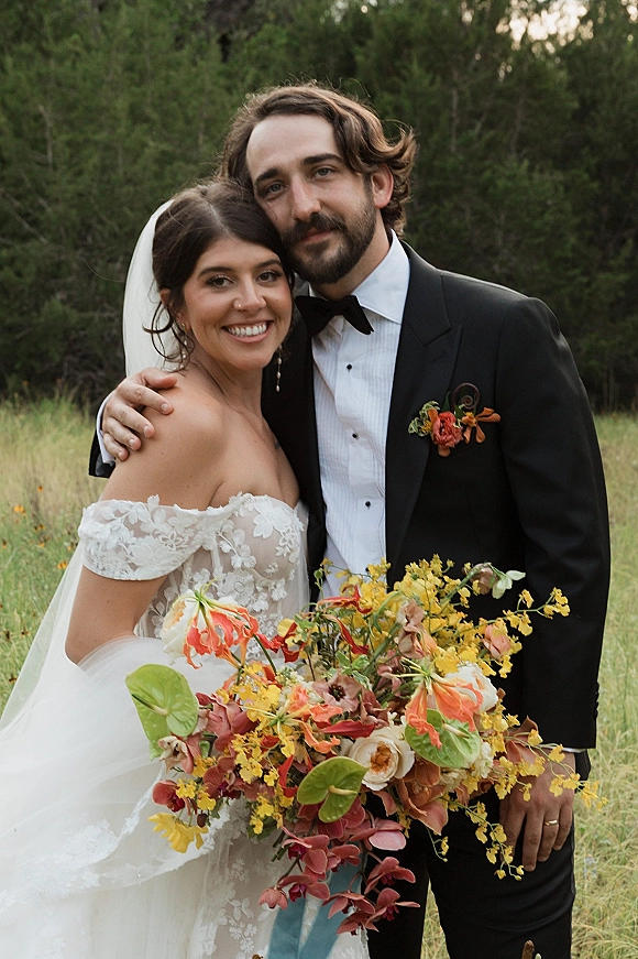 Couple portrait of bride and groom hugging, bride in off-shoulder lace dress with veil and orange-yellow bouquet in a meadow of wildflowers