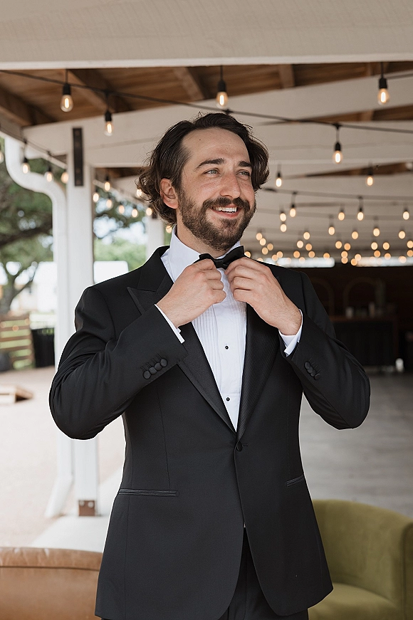 Groom portrait of a bearded man adjusting his bow tie in a black tuxedo with boutonniere under a covered patio with string lights