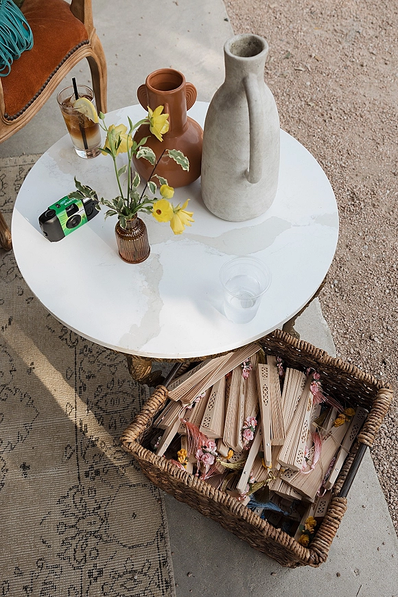 Wedding welcome table with yellow flower bud vases, wicker basket of handheld fans, iced drink cups, and film camera on a patio