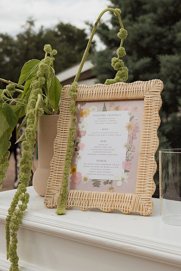 Wedding bar menu in a rattan frame with a floral border, set on a white outdoor bar with greenery and beige vase under trees