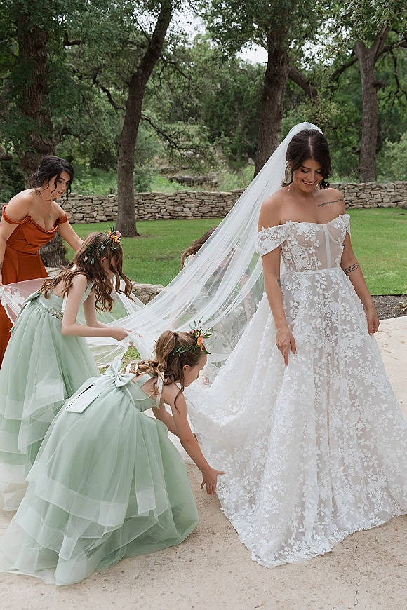 Bridal party moment as bride with flower girls holds her long veil while a bridesmaid adjusts her off-shoulder lace gown in a garden by a stone wall