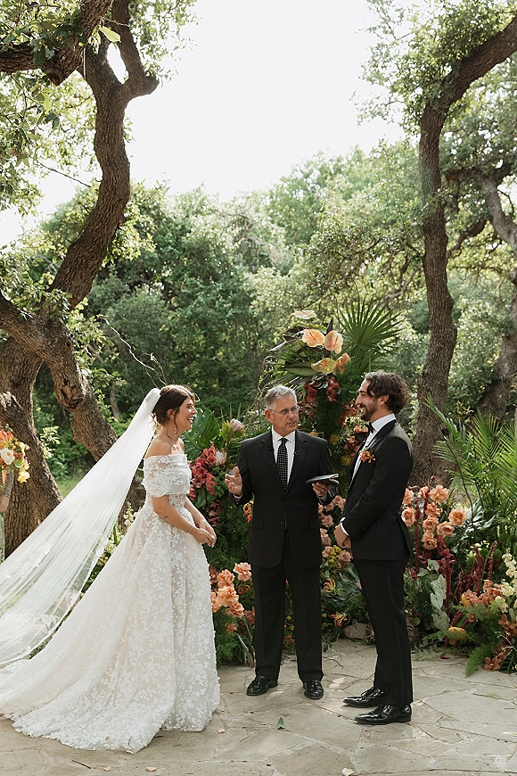 Wedding vows as bride and groom face each other at an outdoor wedding ceremony on a stone patio with lush tropical greenery backdrop and long veil