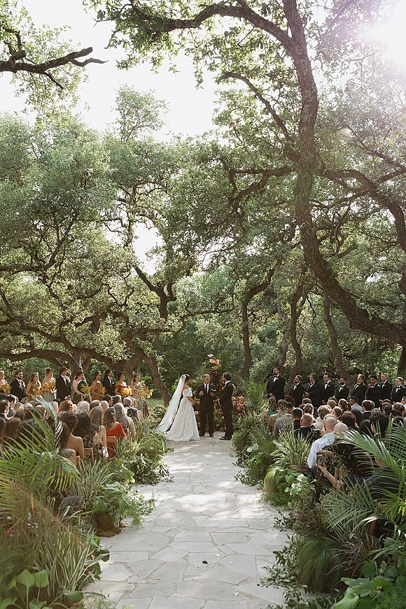 Outdoor wedding ceremony with bride and groom at the altar, floral arrangements lining a stone aisle beneath sunlit trees and guests seated
