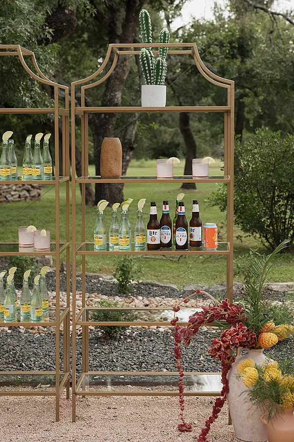 Wedding bar setup with gold shelving, cocktails with lime wedges, beer and canned drinks beside cactus and floral urn on an outdoor lawn