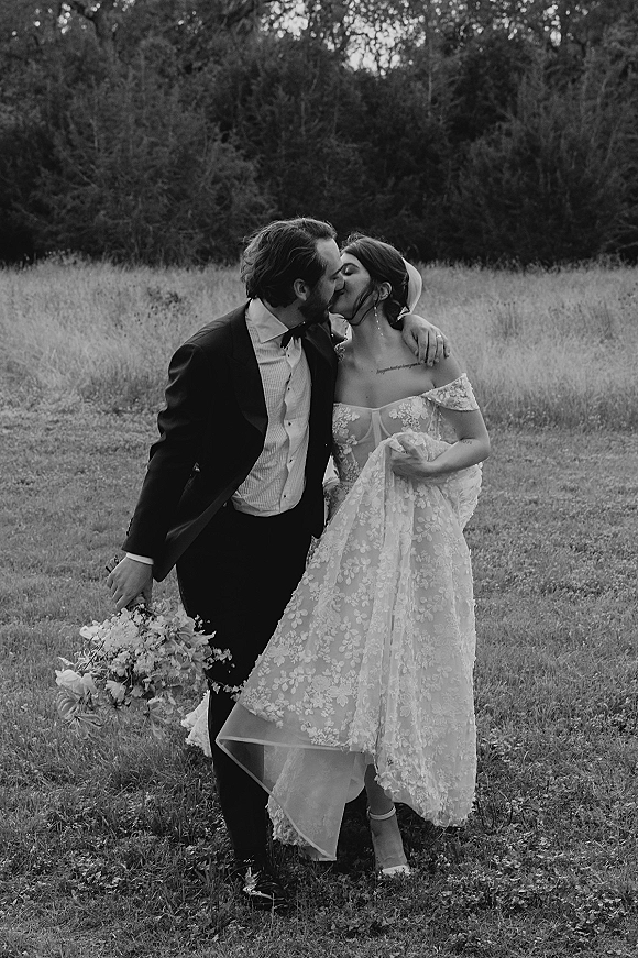 Wedding kiss portrait of bride and groom kissing in a grass field, her lace off-the-shoulder gown and veil flowing, bouquet in hand