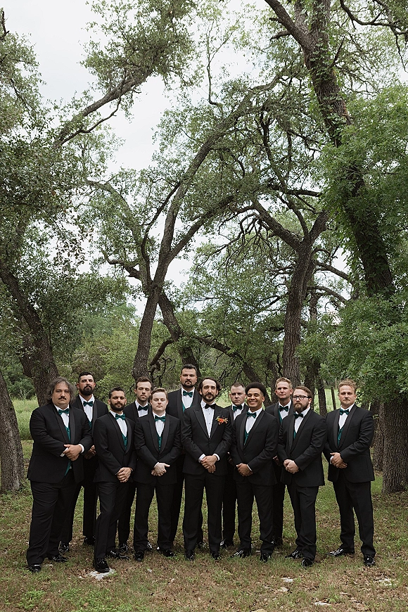 Groomsmen portrait of the groom with groomsmen in black tuxedos and teal vests, standing on grass with trees in a woodland setting
