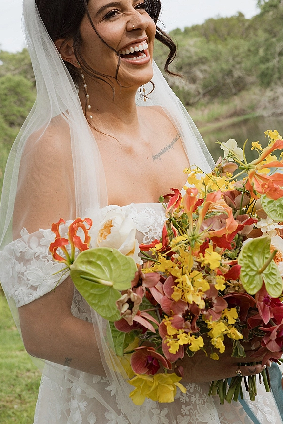 Bridal portrait of a bride laughing, holding a tropical bridal bouquet in an off-the-shoulder lace dress with veil on a green lawn