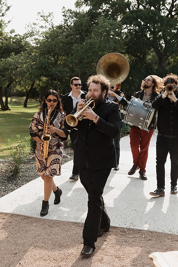Wedding band of brass musicians in black suits, one wearing sunglasses, marching along an outdoor lawn walkway with trees behind