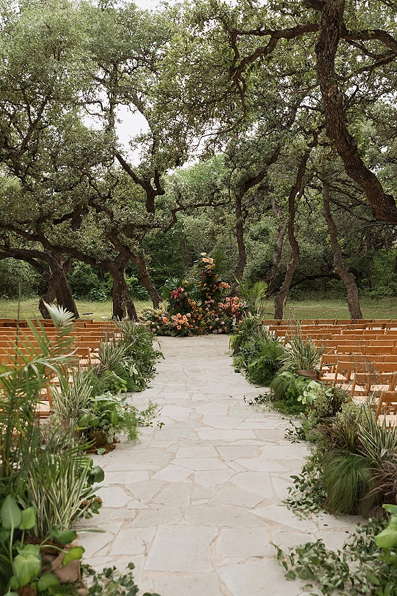 Outdoor ceremony setup with a stone aisle lined in greenery and wooden folding chairs beneath oak trees in a forest clearing