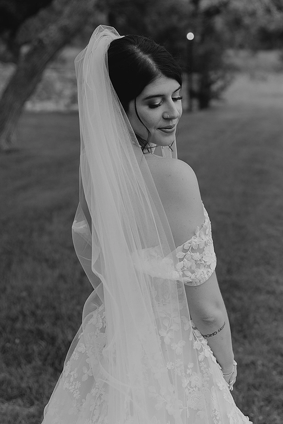 Bridal portrait in black and white of a bride looking over her shoulder, long veil flowing over a lace off-shoulder gown on a garden lawn