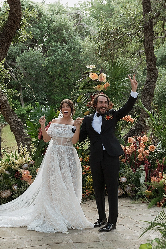 Wedding couple portrait of bride and groom cheering on a stone patio, her strapless lace dress and tropical anthurium accent in garden greenery