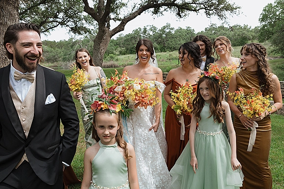 Wedding party portrait with bridal party laughing as the bride holds a bouquet, gathered on a grassy lawn under oak trees
