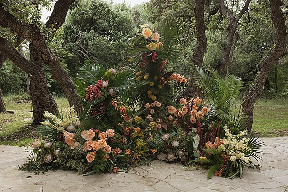 Ceremony floral installation with a wedding floral arch alternative of palm fronds and peach blooms on a stone patio under oak trees