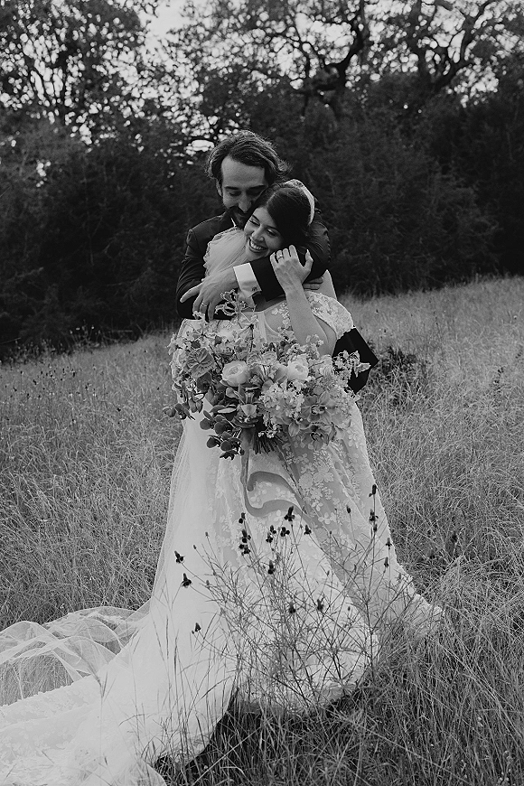 Couple portrait of bride and groom embrace, her lace wedding dress and bouquet in a wildflower meadow under overcast sky