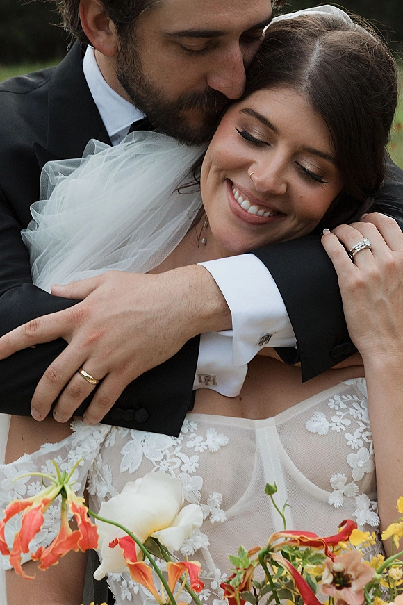 Couple portrait of bride and groom embrace, forehead kiss beside outdoor greenery, with bridal veil and lace dress details visible