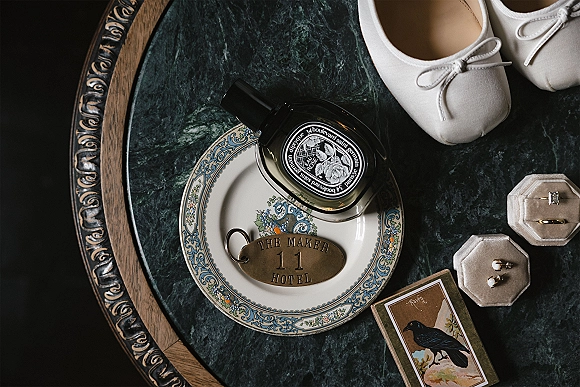 Wedding details flatlay with bridal perfume flatlay featuring rings in velvet boxes, earrings, white flats, and hotel key fob on green marble