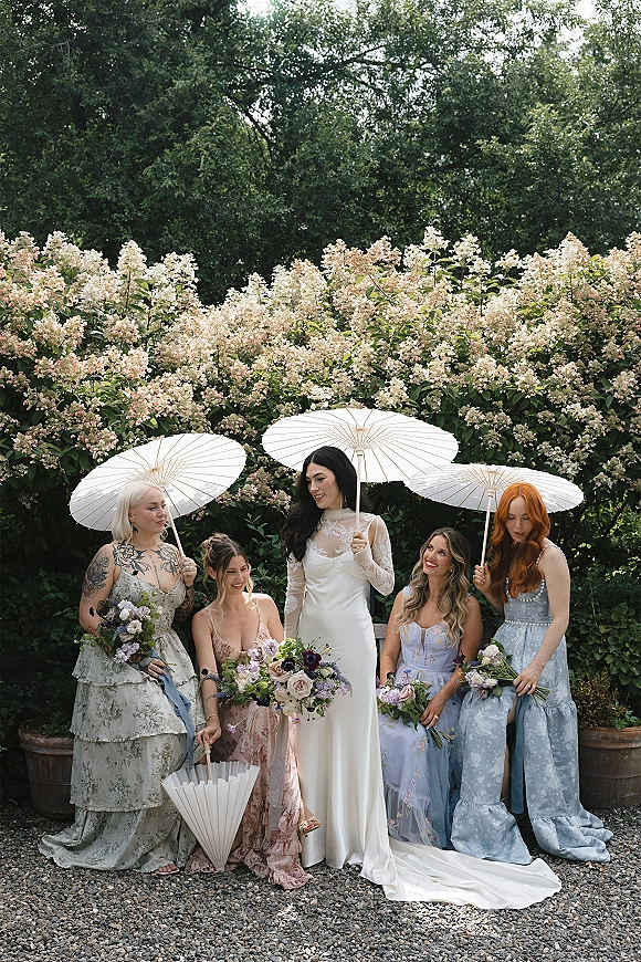 Bridal party portrait with bride with bridesmaids holding white parasols and bouquets on a gravel path beside a flowering hedge