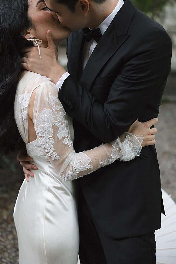 Wedding kiss as groom in black tuxedo cups bride’s face, lace long sleeves and pearl drop earrings visible, greenery behind on gravel path