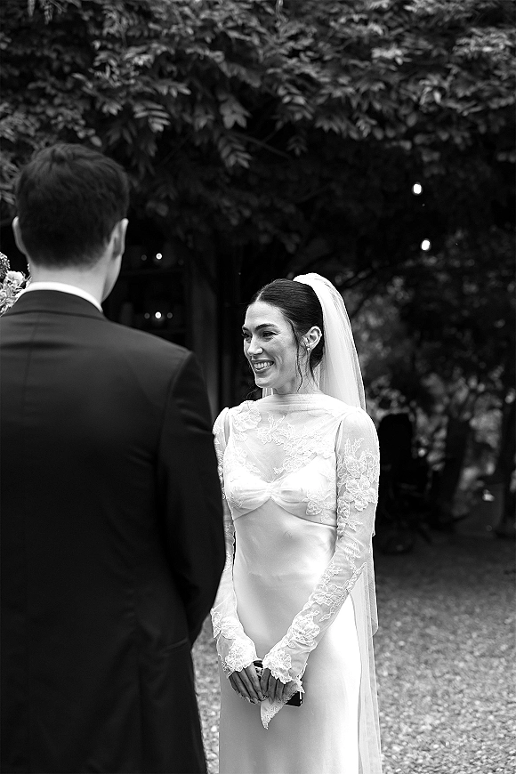 Wedding ceremony moment as bride smiles at groom, lace-sleeved dress and veil under string lights along a garden gravel path