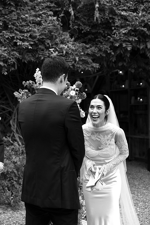 First look moment as groom sees bride in lace-sleeved dress and veil, holding bouquet and vows booklet on a garden path under string lights