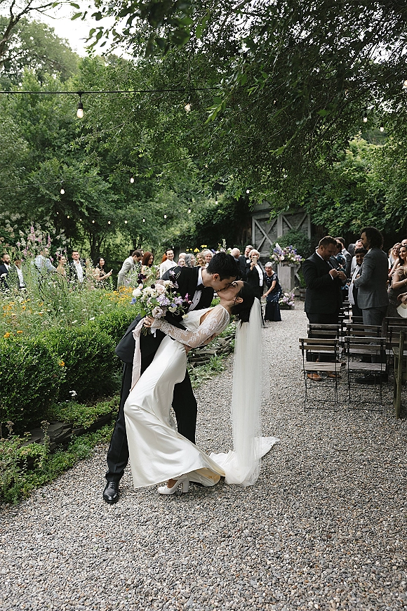 Wedding kiss portrait of bride and groom in a dip kiss, her veil flowing and bouquet raised, as guests cheer on a gravel garden aisle under string lights