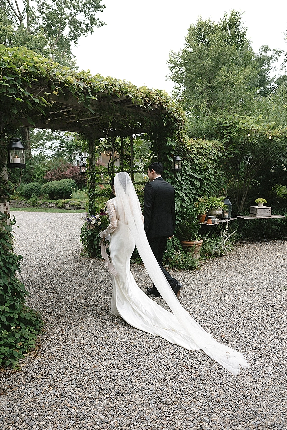 Wedding couple portrait of bride and groom walking away under a greenery pergola, bride’s long veil trailing as she holds a bouquet on a gravel path