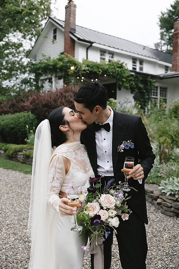 Wedding kiss portrait of bride and groom kissing, holding champagne flutes, with her veil and bouquet on a garden path under string lights