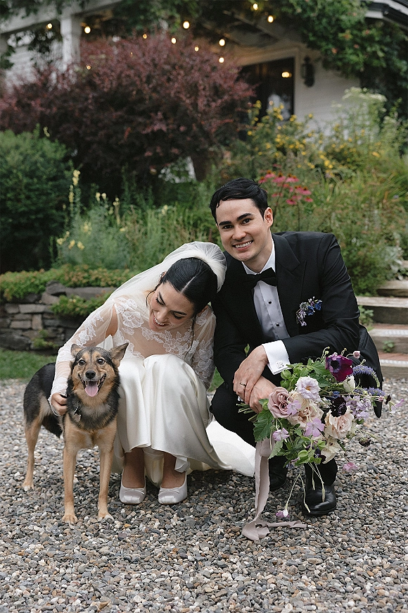 Couple portrait with bride and groom crouching to pet their dog, groom holding bouquet, string lights glowing over garden steps