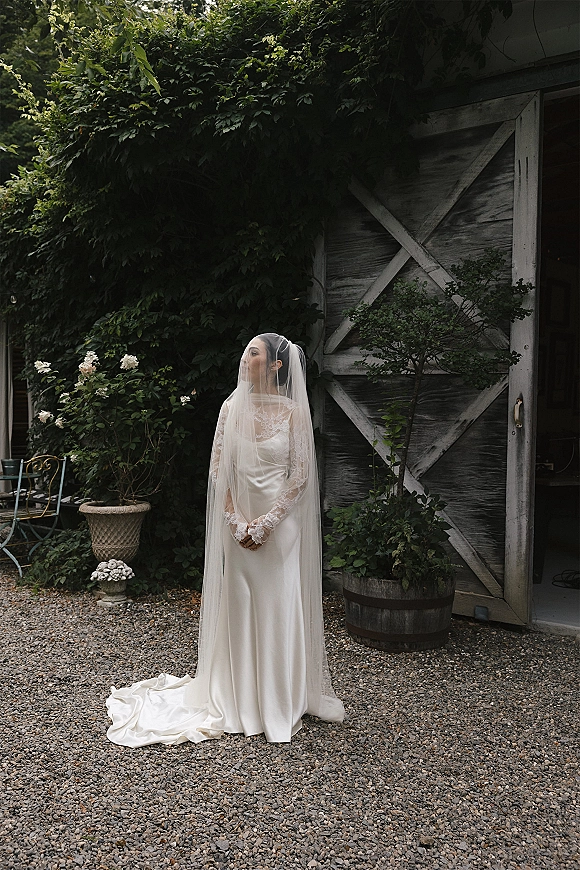 Bridal portrait of a bride with veil over face and lace sleeves, long train trailing by an ivy-covered wall and rustic barn door outdoors