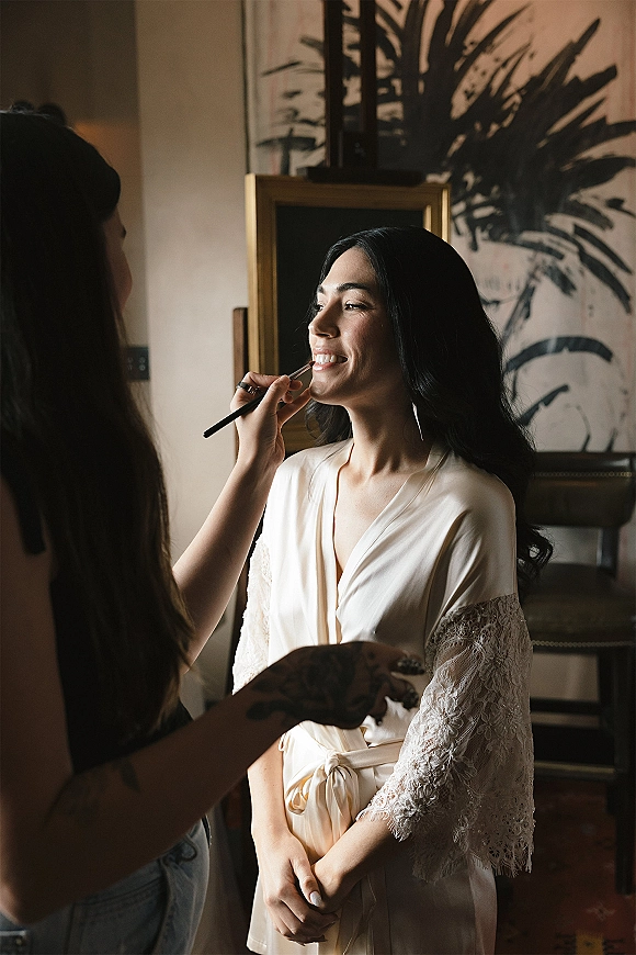 Bridal makeup as a wedding makeup artist applies lip color to a bride in a lace robe with drop earrings in an indoor room