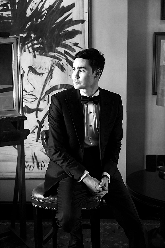 Groom portrait in a black tuxedo groom look, seated on a leather stool with bow tie and cufflinks, lit by window light near framed art