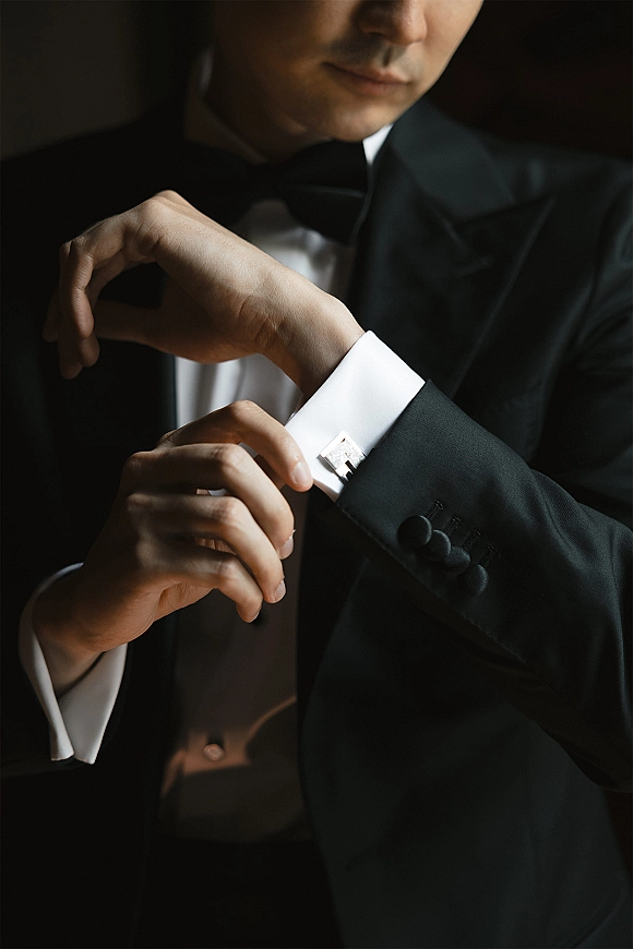 Groom getting ready, adjusting cufflinks on a white French cuff shirt beneath a black tuxedo jacket and bow tie in soft shadows