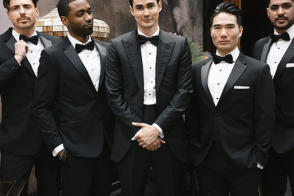 Groomsmen portrait in black tie wedding party style, standing in black tuxedos and bow ties beside a dark wall with greenery