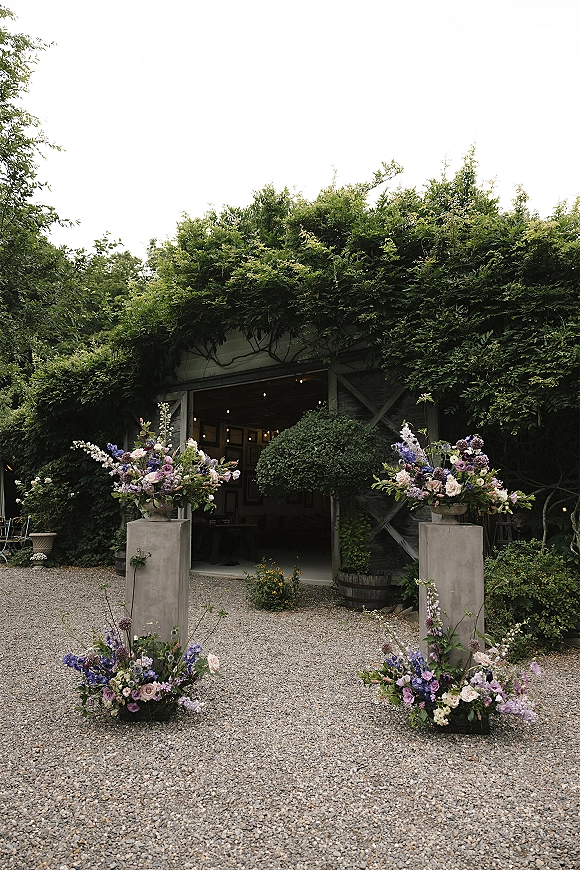 Ceremony entrance decor with wedding entrance flowers on tall pedestal stands and urns, framed by ivy-covered structure and string lights