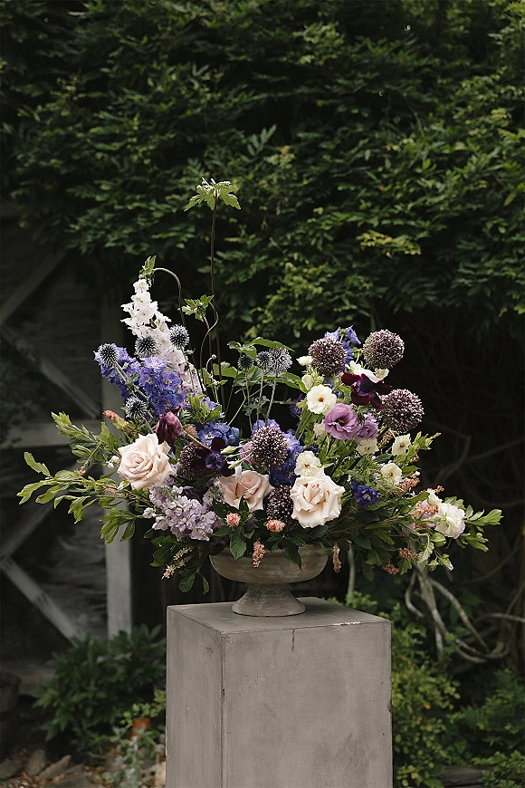 Wedding floral arrangement in a stone urn with roses and purple-blue blooms, lush greenery, set on a pedestal among evergreens