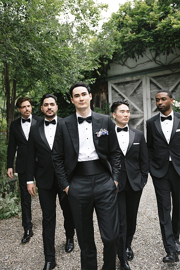 Groomsmen portrait of men in black tuxedos with bow ties and boutonnieres walking on a gravel path by ivy barn doors