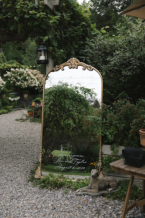 Wedding welcome sign on an ornate gold mirror with calligraphy lettering, lantern, and string lights along a garden gravel path under a pergola