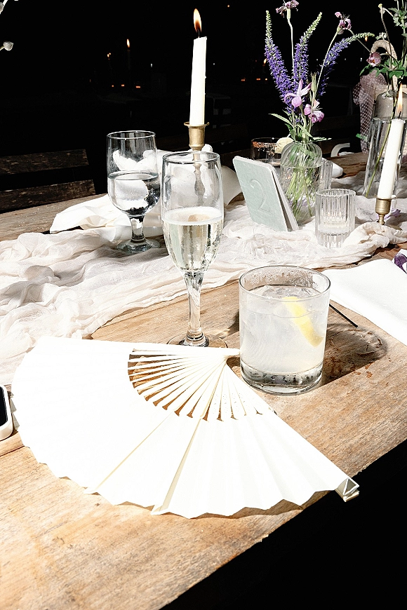 Reception tablescape with a gauze wedding table runner, wildflower bud vases and taper candles in brass candlesticks on a rustic wood table in a dark room