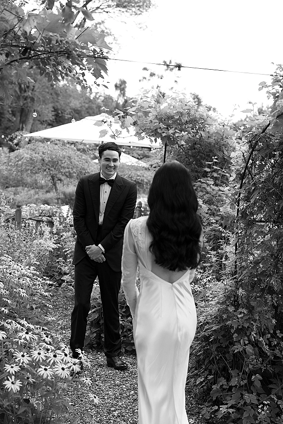 Wedding first look as bride walks toward groom in black tuxedo with bow tie, string lights above, garden path and trees behind