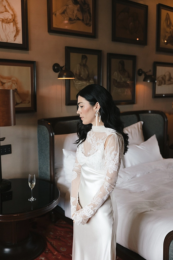 Bridal portrait of a getting ready bride in a long sleeve lace gown holding a champagne flute in a hotel bedroom with bedside lamps