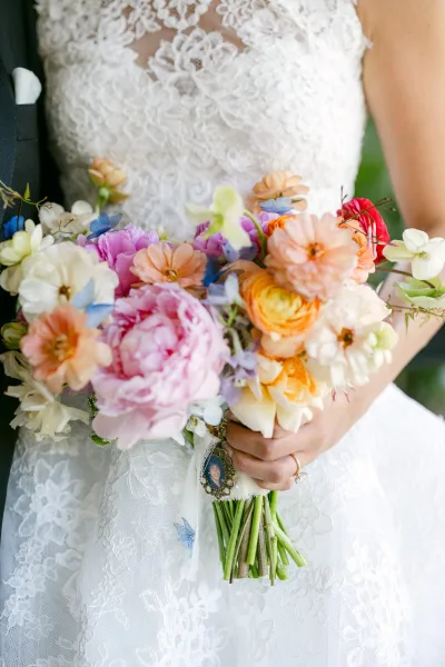 Bridal bouquet of pastel peonies, ranunculus, and zinnias, colorful wedding bouquet with ribbon and photo charm against blurred greenery