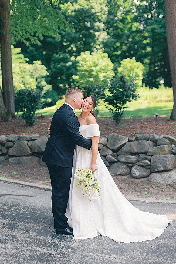 Couple portrait of groom kissing bride’s cheek as she hugs him, holding a bridal bouquet in front of a stone wall and garden greenery