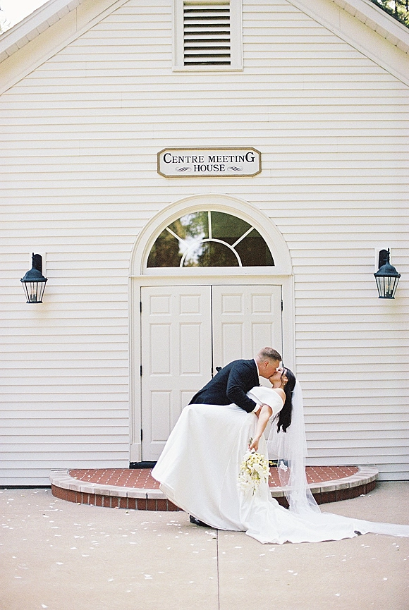Wedding kiss portrait of groom dipping the bride as they kiss, her long veil flowing and bouquet held at a white chapel entrance