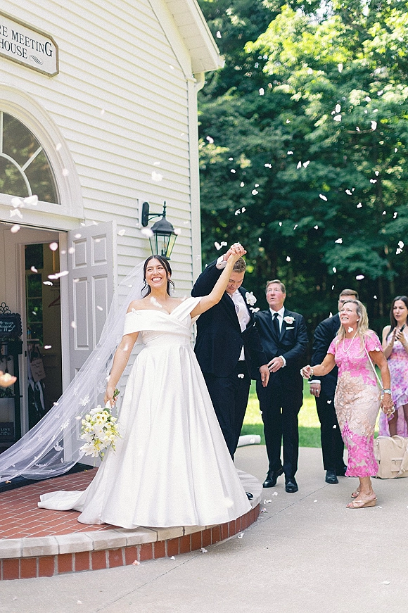 Wedding recessional as newlyweds leave the chapel, bride in long veil with bouquet and groom in tux amid flower petals on brick steps
