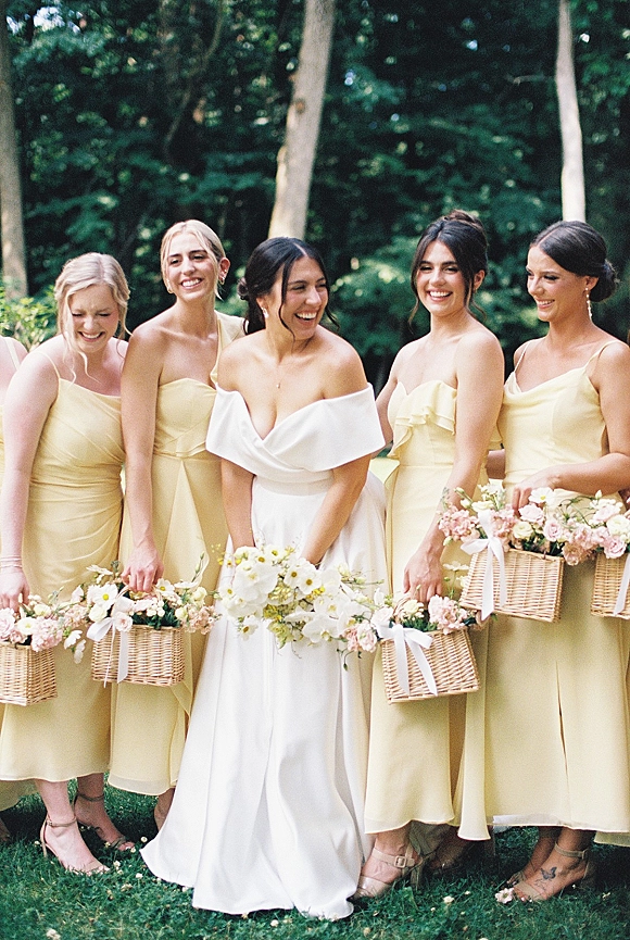 Bride with bridesmaids in satin dresses holding wicker flower baskets with pastel blooms and white ribbons on a garden lawn with trees