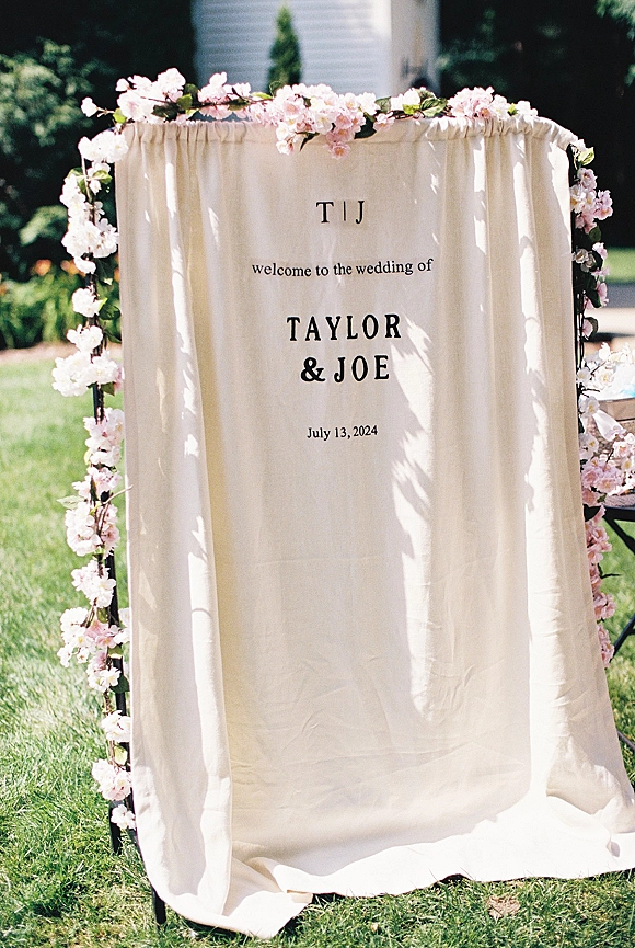 Wedding welcome sign on a metal frame with a fabric drape and flower garland, displayed on a grass lawn by a house in daylight