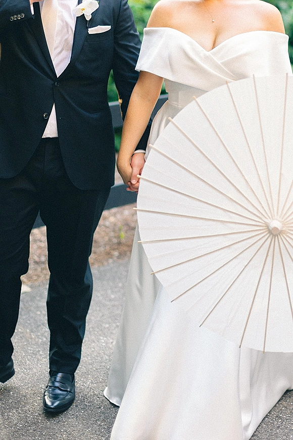 Couple portrait of bride and groom holding hands, bride in off the shoulder wedding dress with paper parasol on a green path