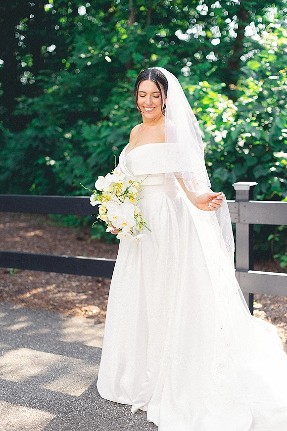 Bridal portrait of a smiling bride holding bouquet in a strapless satin wedding dress and long veil on a paved path by trees and a wooden fence