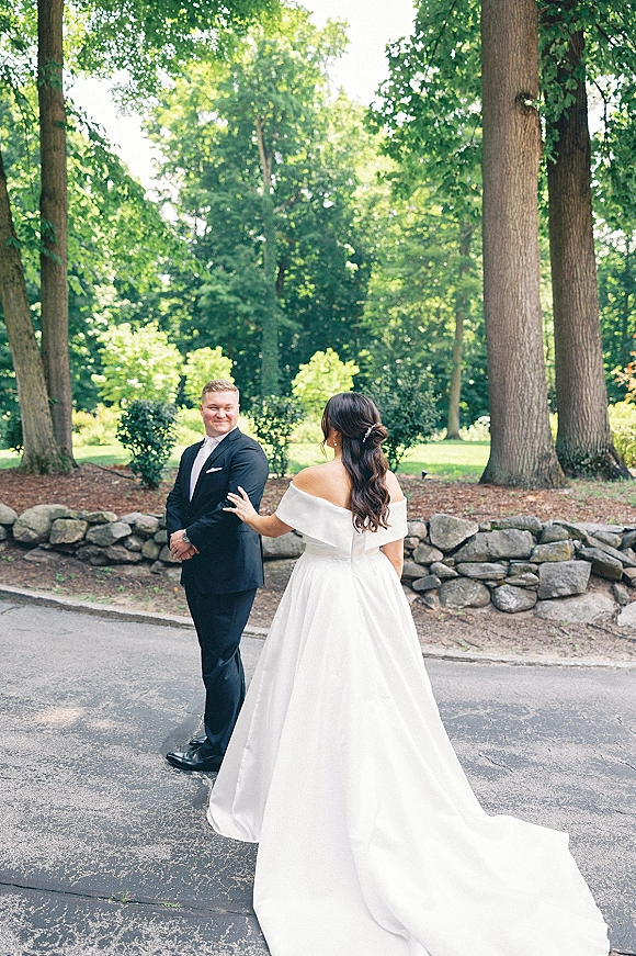 First look moment as bride taps groom’s shoulder, her off-the-shoulder gown with long train flowing on a tree-lined park path by stone wall