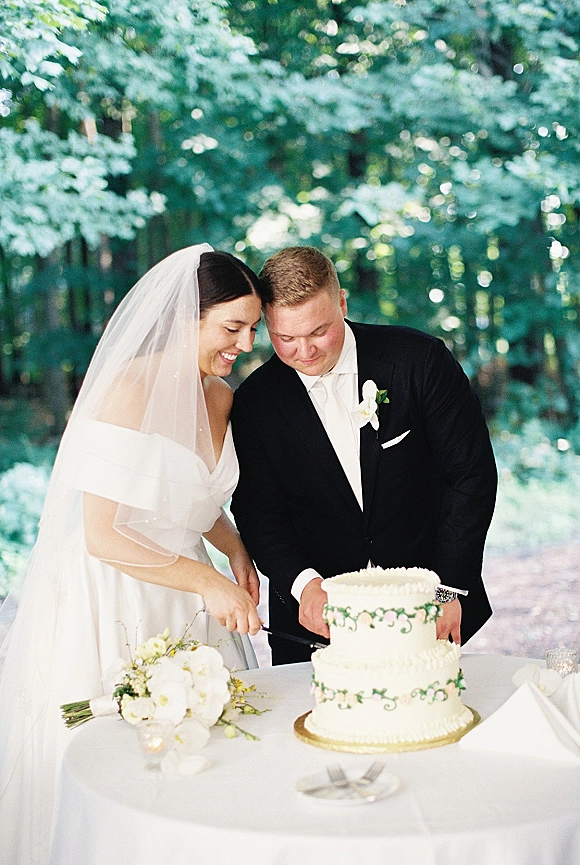Wedding cake cutting with bride and groom cutting cake beside a two-tier white cake, bouquet and knife on table in outdoor greenery
